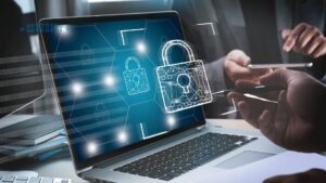 Na zdjęciu widzimy laptopa z wyświetlonym obrazem symbolizującym bezpieczeństwo cyfrowe, przedstawionym w postaci zamka. W tle widać osoby dyskutujące lub pracujące, jedna z nich trzyma tablet. Obraz sugeruje tematykę związaną z ochroną danych i cyberbezpieczeństwem, w kontekście biznesowym lub biurowym.