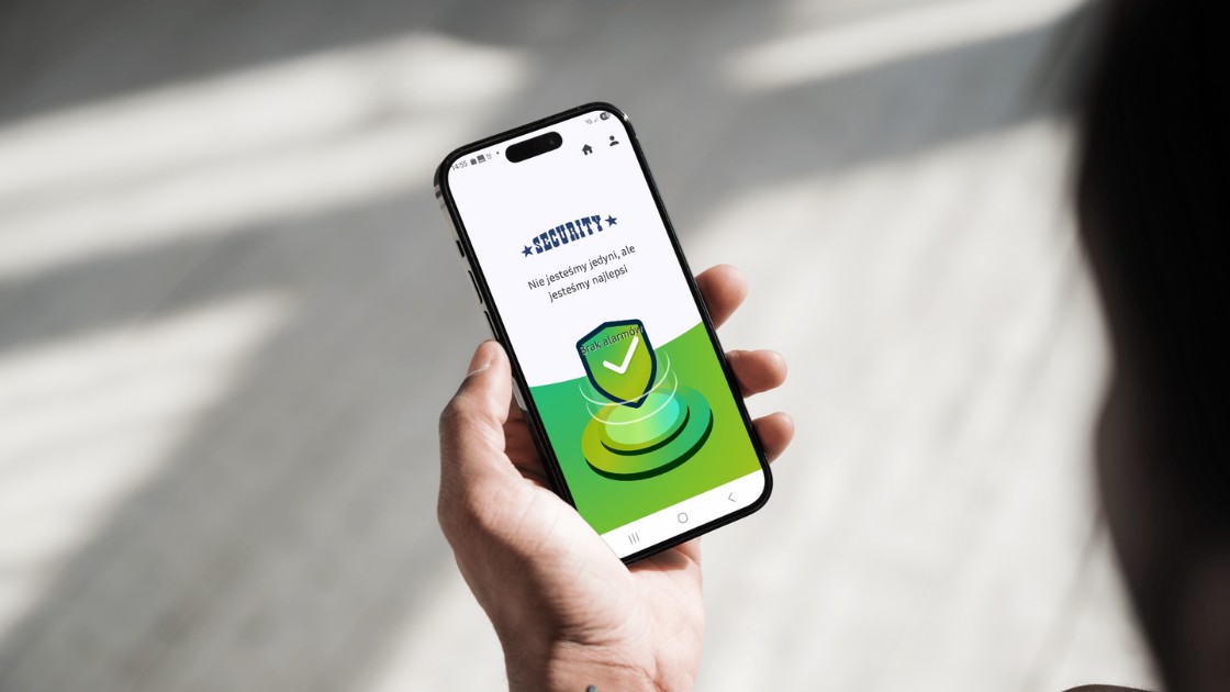 Osoba trzymająca smartfon z uruchomioną aplikacją Monpatrol, na ekranie widoczny komunikat "Brak alarmu" i logo SECURITY.